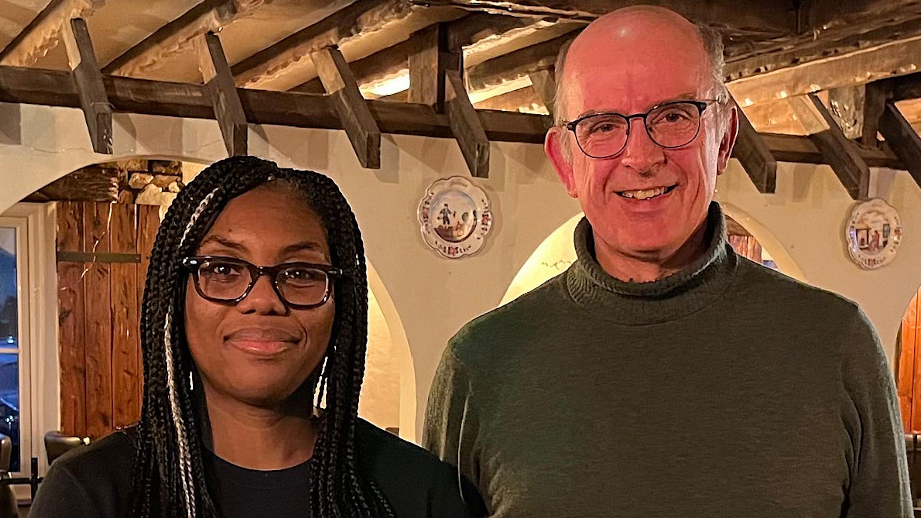 A head and shoulders photo of Kemi Badenoch who stands on the left wearing thick-rimmed glasses, beside Andrew Coles in a green turtle neck jumper.