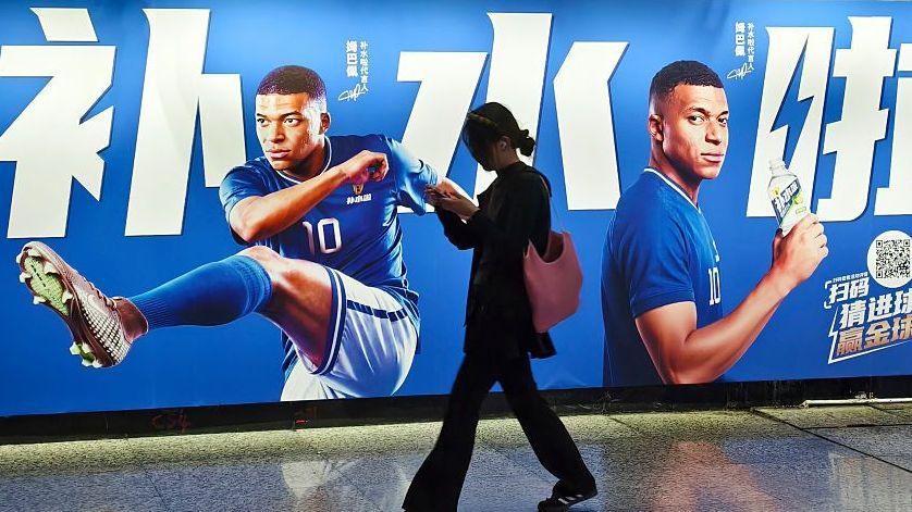 Pedestrians pass by an electrolyte drink advertisement for French football player Kylian Mbappé in the subway in Shanghai, China on June 30, 2026