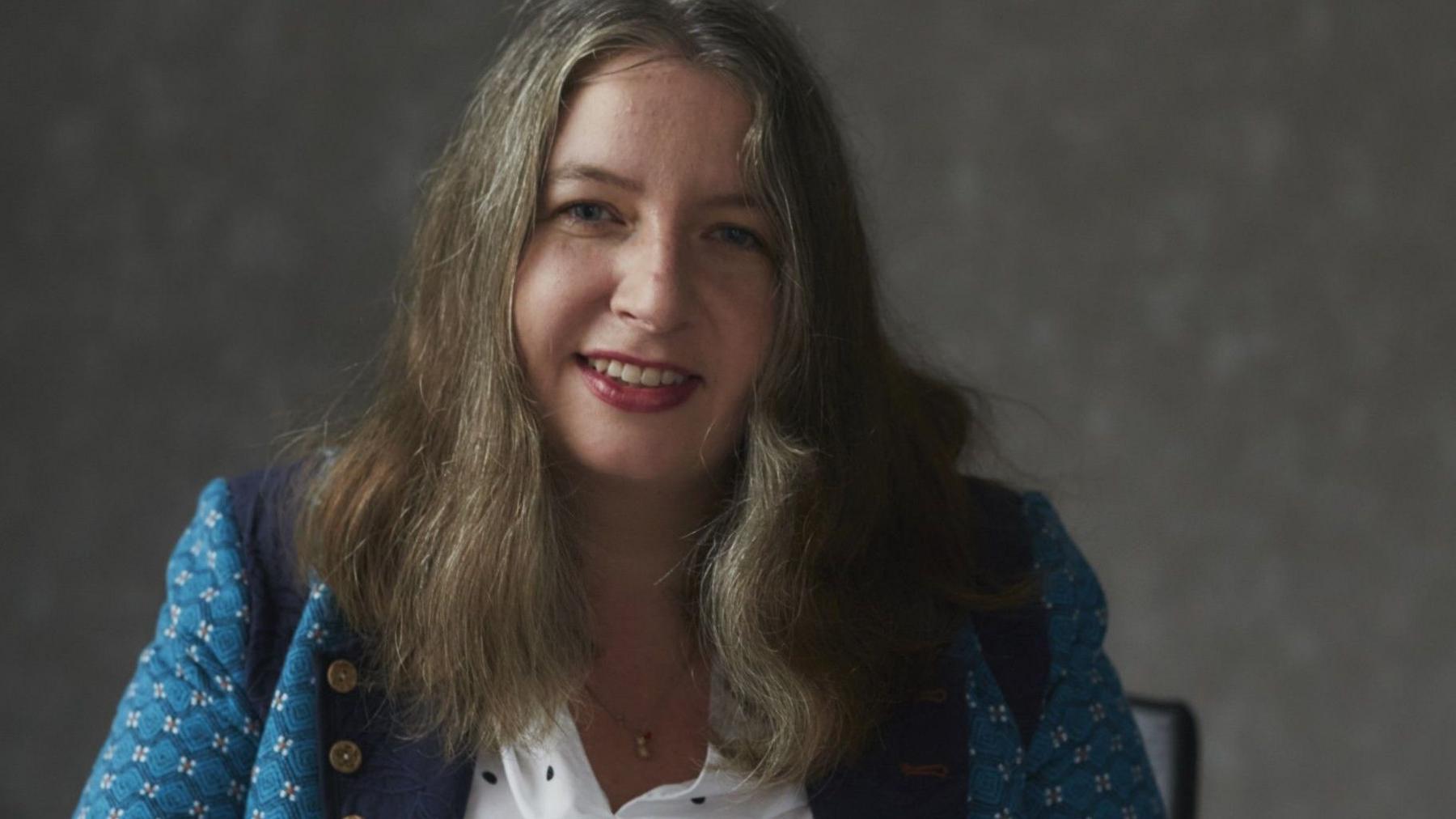A woman with long brown and grey hair smiling. She is sitting at a table with a grey wall in the background. She is wearing a white shirt and a black jacket with white flowers on it