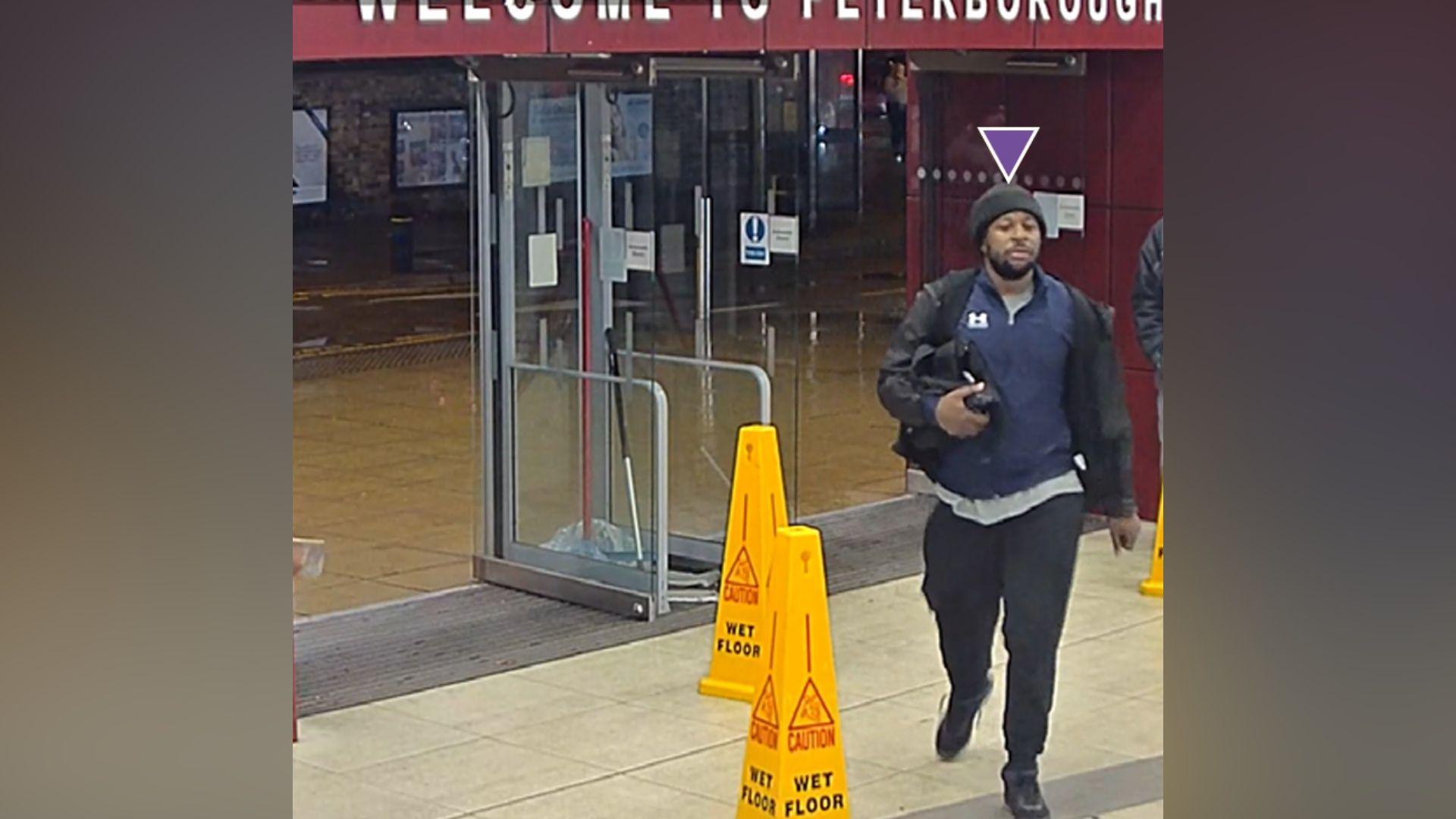 A man in dark clothing and black woolly hat carries a small black bag under his right arm. He is outside glass doors at a railway station and is walking past yellow cones that say "CAUTION WET FLOOR" on them.