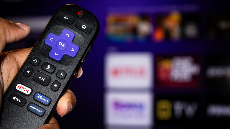 A Roku remote with the Roku OS app grip hazy in the background.