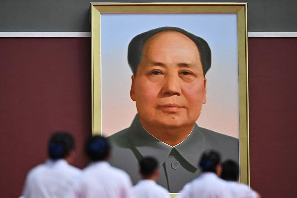Large framed portrait of Mao Zedong hanging on a red wall, viewed from below with several people in light-coloured clothing standing and facing the portrait in the foreground.